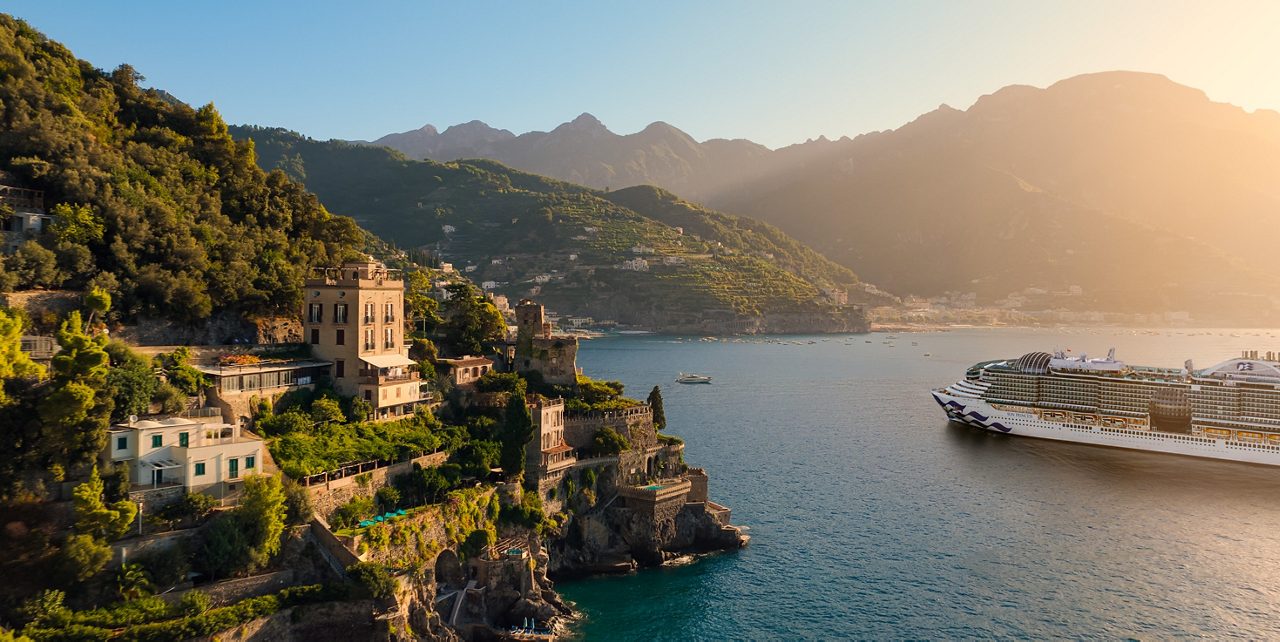 A cruise ship gliding through a narrow bay in Kotor, Montenegro, surrounded by steep green mountains, red‑roofed coastal towns, and bright blue water under a clear sky.